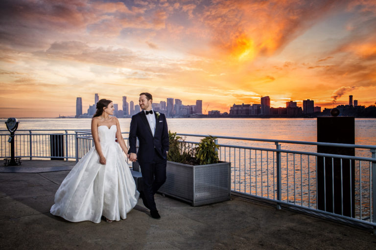 Bride and Groom walking on The Lighthouse veranda, the Hudson river, New Jersey and the Sunset are in the background