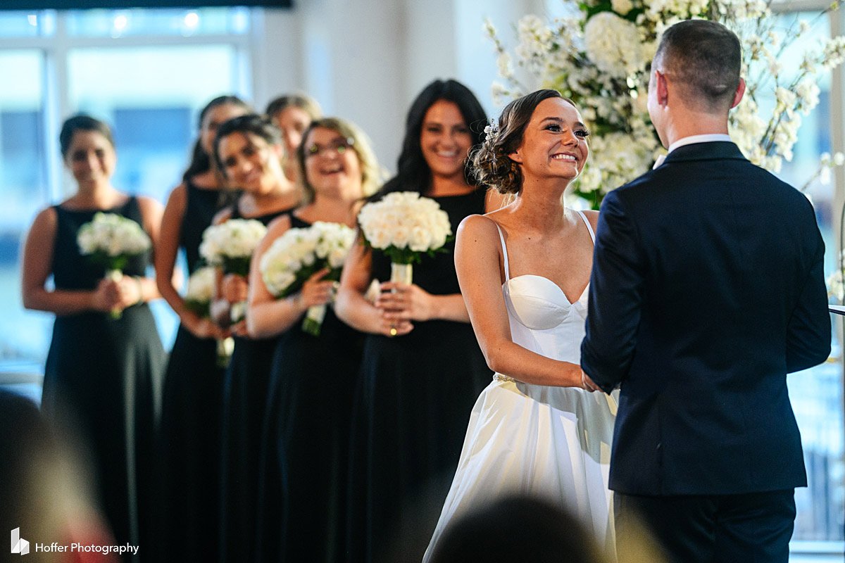 Bride standing at the ceremony, holding the groom's hands, she is smiling, and bridesmaids are behind her