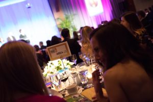 Guests seated at their dinner table listening to the speaker on stage at Pier Sixty
