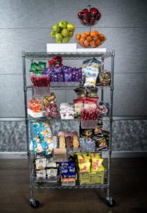 A rolling cart filled with different types of snacks including fruits, candy, chips, and cookies
